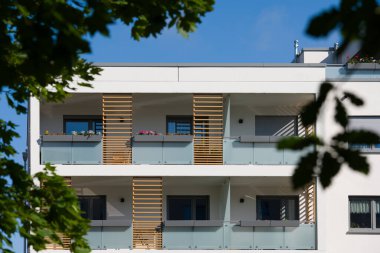 Güneşli bir günde modern bir apartmanın dışında. Ön cephede buzlu cam korkuluklar ve dekoratif ahşap çıtalarla çağdaş balkonlar yer alıyor. Önplandaki yeşil bir ağaç şehir ortamına doğanın bir dokunuşunu ekler. Modern için kavram