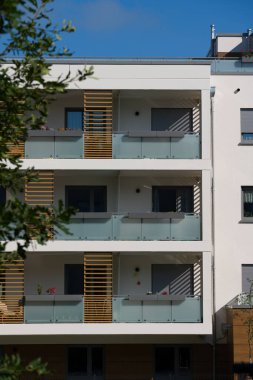 Güneşli bir günde modern bir apartmanın dışında. Ön cephede buzlu cam korkuluklar ve dekoratif ahşap çıtalarla çağdaş balkonlar yer alıyor. Önplandaki yeşil bir ağaç şehir ortamına doğanın bir dokunuşunu ekler. Modern için kavram