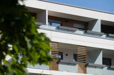 Güneşli bir günde modern bir apartmanın dışında. Ön cephede buzlu cam korkuluklar ve dekoratif ahşap çıtalarla çağdaş balkonlar yer alıyor. Önplandaki yeşil bir ağaç şehir ortamına doğanın bir dokunuşunu ekler. Modern için kavram