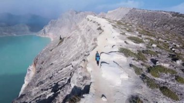 Aktif bir volkan dağı ve sisle kaplanmış turkuaz sülfür gölü olan bir kayanın üzerindeki bir adamın güzel bir görüntüsü.