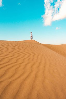 Beyaz bir abaya giymiş genç adam Sahara çölünde bir kum tepesinin kenarında duruyor.