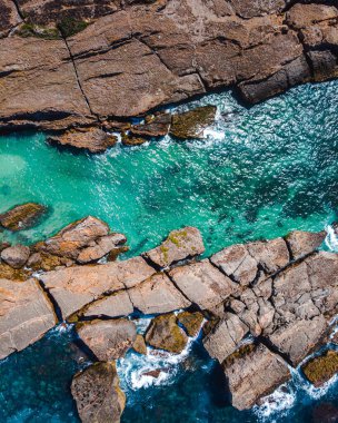 Rocky kıyı şeridinin hava görüntüsü çarpışan turkuaz dalgalarla karşılaşıyor, dinamik bir deniz ve taş çarpışması.