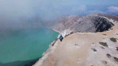 Kawah Ijen volkanı, Doğu Java, Endonezya 'da kayalıklarla çevrili turkuaz sülfür gölü manzaralı bir dağın tepesinde duran adam..