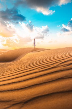 Geleneksel Abaya kıyafetli genç adam, dalgalanan dokusuyla ve gün batımında Dubai çöl kumunda, Dubai Emirlikleri 'nde altın rengi ile güzel çölde duruyor.