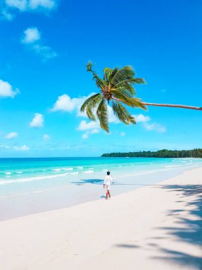 Tatilde olan bir adam, Hindistan cevizi palmiyeleri ve turkuaz deniz suyu manzaralı tropik bir cennetin sahilinde yürüyor..