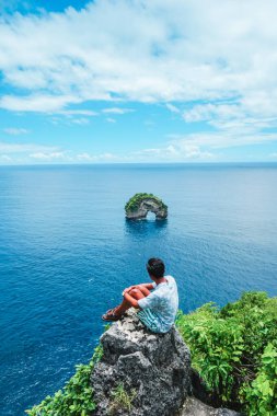 Uçurumun kenarında oturan bir adam güzel bir deniz ve ada manzaralı.