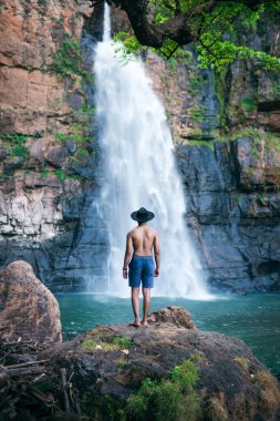 Arka planında büyük bir şelale olan bir kayanın üzerinde duran adam. Seyahat ve tatilin doğa manzarası