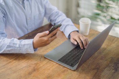 Genç bir adam, girişimci, serbest çalışan, kahve masasındaki koltukta oturuyor, akıllı telefon kullanıyor, dizüstü bilgisayarla çalışıyor.. 