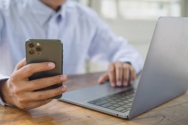 İş adamı cep telefonu kullanıyor ve ofisteki dizüstü bilgisayarda çalışıyor. Çevrimiçi çalışan bir adam, kafede internette sörf yapan, mesai yapan, tele-iş yapan, uzaktan çalışan, telekomünikasyon yapan.