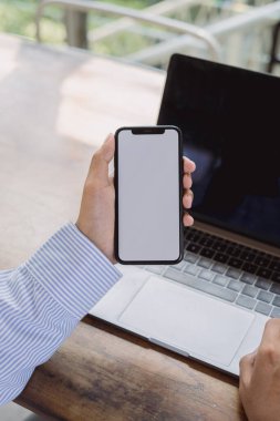 Siyah cep telefonlu, boş beyaz ekranlı ve ahşap masadaki laptoplu ellerin görüntüsü.