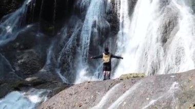 Genç erkek turist dik taş duvarın önünde duruyor ve şelale manzarasının tadını çıkarıyor. Çevre, su dolaşımı, turizm kavramı.