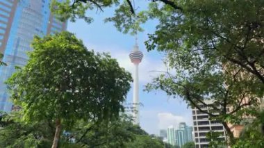 Menara Kuala Lumpur (KL Tower) ön planda yemyeşil ağaçlar ve arka planda açık mavi gökyüzü ile. İkonik telekomünikasyon kulesi ve Malezya 'da popüler turizm merkezi.