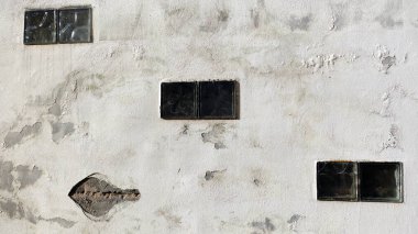 Eski bir duvarın yüzeyinde, çökmekte olan yapısına gömülmüş üç cam bloğu gösteren bozulma ve ihmal belirtileri var. Duvarlar desenli yüzey boyaların soyulduğu bölgelerle işaretlenmiş.