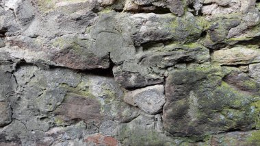 Yakın çekim, yıpranmış bir taş duvarın karmaşık dokularını ve desenlerini ortaya çıkarır. Yüzey, çatlaklarda büyüyen yeşil yosun parçalarıyla vurgulanan gri ve kahverengi tonlarıyla işaretlenmiştir. Taşlar yaşlılık belirtileri gösteriyor, smo ile