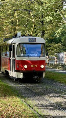 Prag 'daki kırmızı-beyaz tramvayın dikey fotoğrafı, sonbahar ağaçlarının arka planına çizilmiş. Tarihsel cazibeyi, mevsimsel renkleri ve sonbaharda klasik Çek şehir manzarasını yakalar