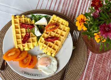 Yumurtalı tuzlu waffle, domates ve bir tabakta otlar. Masanın üzerinde bir tabak waffle ve çiçek olan samimi bir kahvaltı sahnesi. Sağlıklı bir kahvaltı ve dengeli bir yaşam tarzının mükemmel bir örneği.