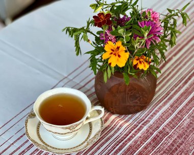 Bir vazo çiçeğin yanındaki masaya yerleştirilmiş porselen çay. Samimi ve zarif bir sahne, çay saatini, rahatlamayı ya da huzurlu bir sabahı resmetmek için ideal.