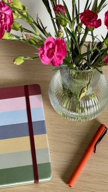 Çalışma ya da çalışma alanının dikey fotoğrafı. Kapalı bir defter, kalem ve bir buket çiçek, rahat ve üretken bir atmosfer yaratıyor. Ofis, eğitim ya da ev dekorasyonu için ideal.