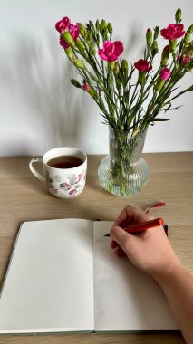 Bir kadının elinde boş sayfalarla dolu bir defterin üstünde dolmakalem tutarken bir kupa çay ve bir vazo çiçekle bir masanın üzerinde dikey fotoğrafı. Yaratıcılık, günlük ve iş temaları için mükemmel.