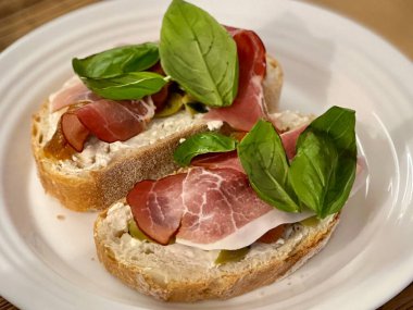 Beyaz tabakta jambonlu ve fesleğenli iki sandviç. Yemek, gurme, öğle yemeği ve mutfak teması için ideal. Taze malzemeler ve lezzetli yemekler sergilemek için mükemmel..