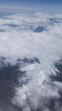 Aşağıdaki dağları ve bulutları gösteren bir uçak penceresinin dikey görüntüsü. Seyahat, hava manzarası, dağ manzarası, havacılık, gökyüzü manzarası ve uçuş perspektifleri için ideal..