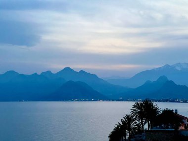 Akdeniz 'de gün batımı, palmiye ve dağ siluetleriyle, ön planda sakin deniz. Yaz seyahati, kıyı manzarası, tatil, doğa, sıcak akşam manzarası, Antalya, Türkiye. 