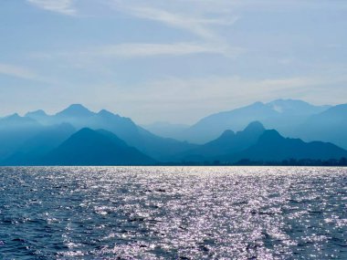 Sakin denizin yukarısındaki dağ silueti ve ışıldayan su. Akdeniz, yaz tatili, seyahat, kıyı manzarası, huzurlu manzara, Antalya, Türkiye, doğa, huzur manzarası, sıcak akşam ışığı. 