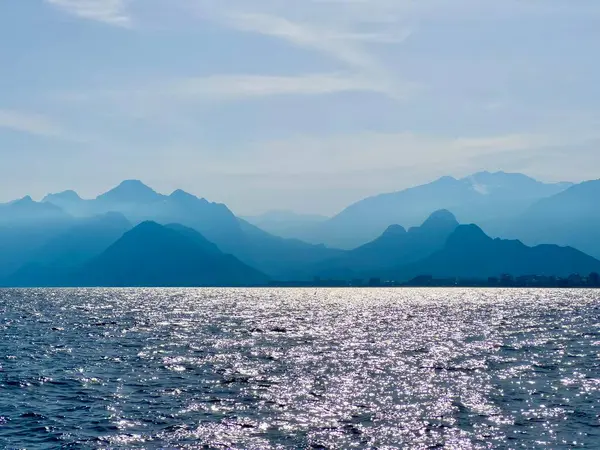 Sakin denizin yukarısındaki dağ silueti ve ışıldayan su. Akdeniz, yaz tatili, seyahat, kıyı manzarası, huzurlu manzara, Antalya, Türkiye, doğa, huzur manzarası, sıcak akşam ışığı. 