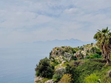 Çiçek açan salkımlı kayalık yamaçlar ve parçalı bulutlu gökyüzü altında sisli dağlara ve denize tepeden bakan tropikal yeşillikler. Bahar manzarası, Akdeniz florası, kıyı bahçesi, doğa, Antalya, Türkiye. 