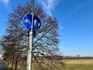 Arka planda arazi, ağaçlar ve gökyüzü olan ayrı yayalar ve bisiklet yollarını gösteren mavi trafik levhası. Açık hava altyapısı, bisiklet, yürüyüş, kırsal bölge, ulaşım, yol güvenliği. 