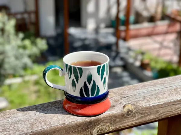 Renkli seramik bardakta bir fincan çay ahşap korkuluklara yerleştirilmiş bulanık bir bahçe ve veranda arka planı ile. Sıcak açık hava anı, sabah çayı, rahatlama, yavaş yaşam, günlük yaşam tarzı, yaz havası.. 