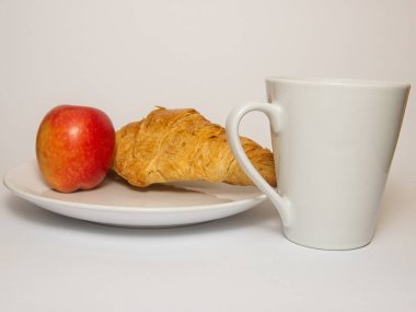 Beyaz bir tabak, kırmızı elma ve kruvasanla birlikte beyaz bir kupa. Düzenek basit bir kahvaltı ya da atıştırmalık öneriyor.