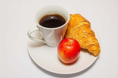 Görüntü, kruvasan, kırmızı elma ve beyaz tabakta bir fincan sade kahveyle kahvaltı setini gösteriyor. Kruvasan altın rengi kahverengi ve pul pul, elma parlak ve kırmızı, kahve koyu ve buharlı.. 