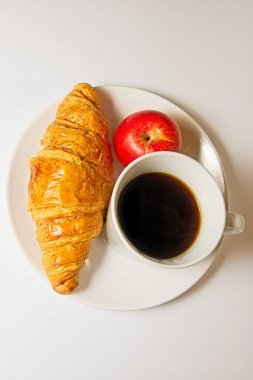 Görüntü, kruvasan, kırmızı elma ve beyaz tabakta bir fincan sade kahveyle kahvaltı setini gösteriyor. Kruvasan altın rengi kahverengi ve pul pul, elma parlak ve kırmızı, kahve koyu ve buharlı.. 