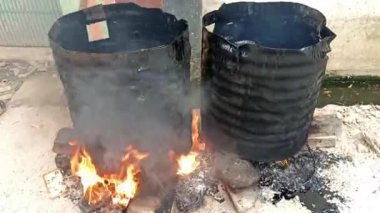 İki büyük siyah varil, yol kömürü katranıyla dolu bir odun ateşinin üzerinde oturur. Dekor açık havada, muhtemelen kırsal ya da yol inşaatı alanında..