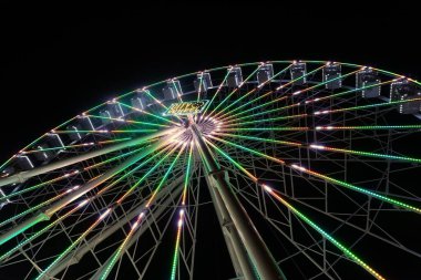 Aşağıdan yukarıya doğru bir dönme dolabın yakın plan fotoğrafı. Tekerlek farklı ışıklarla parlıyordu, kamaralar karanlık akşam gökyüzüne karşı beyazla aydınlanıyordu..