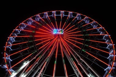 Aşağıdan yukarıya doğru bir dönme dolabın yakın plan fotoğrafı. Tekerlek farklı ışıklarla parlıyordu, kamaralar karanlık akşam gökyüzüne karşı beyazla aydınlanıyordu..