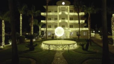 The villa glows with warm light. The palm trees are wrapped in garlands, creating a charming festive atmosphere. In the center of the fountain is a sphere sparkling with bright lights of garlands.