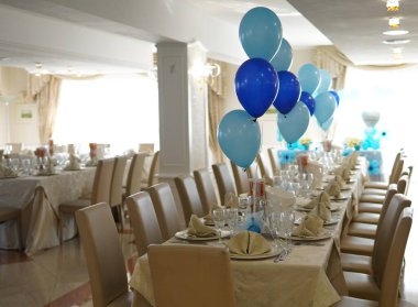 Zarifçe balonlarla şenlik masası kur. Masa krem masa örtüleriyle süslenmiş, sandalyeler döşenmiş. Arka planda beyaz bir piyano ve altın dekoratif elementler var..