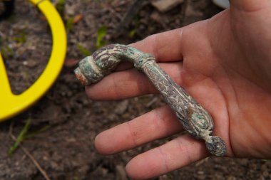 Metal dedektörü. Eski bir eserin yakın çekimi. Karmaşık oymalar uzun zaman önce unutulmuş bir medeniyete işaret ediyor. Kalıntı, topraktan yeni çıkarılmış bir hazine avcısının ellerinde dikkatlice yatıyor..