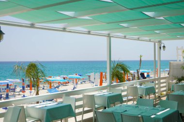 Okyanus manzaralı deniz manzaralı bir restoran. Turkuaz masaları ve beyaz sandalyeleri olan, şemsiyeli güneşli bir plaj ve turkuaz deniz manzaralı, açık deniz manzaralı bir restoran..