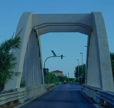 Boş bir yol üzerinde geometrik desenli beton bir kemer köprüsü - açık mavi gökyüzü altında minimalist bir kompozisyon, altyapı veya mimari temalar için ideal.