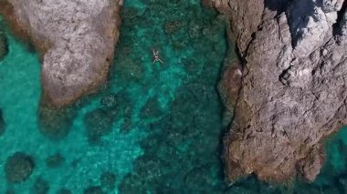 Kayalık kayalıkların arasında denizde yüzen kız, hava manzarası. Kristal berrak mavi su, etkileyici kıyı manzarası, huzurlu yaz tatili, doğal güzellik, insansız hava aracıyla çekilmiş..
