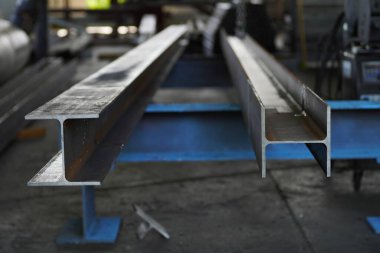 İki uzun paralel çelik I kirişi mavi bir kaynak tezgahının üzerinde duruyor. Görüntü, metalin kaba, endüstriyel dokusunu ve üretim veya inşaat ortamındaki ölçek duygusunu aktarır..