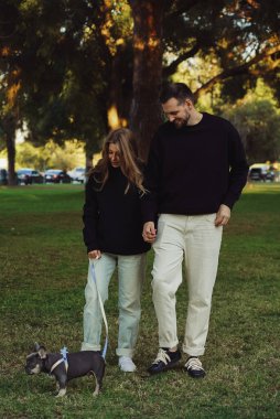 Parkta köpekle gezen bir aile, parkta tasmayla dolaşan bir buldog yavrusu, evin dışında evcil hayvan vakit geçiren genç bir çift.