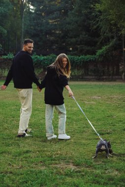 Parkta köpekle gezen bir aile, parkta tasmayla dolaşan bir buldog yavrusu, evin dışında evcil hayvan vakit geçiren genç bir çift.