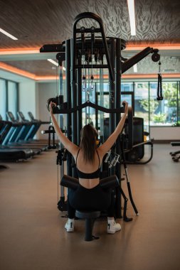 Dikey çekme makinesiyle spor salonunda egzersiz yapan bir kadını yere sermek, bir makinede kol kaslarını şişirmek.