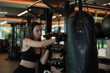 Boks eldivenli bir kadın spor salonunda kum torbasına vurur, kadın antrenman yapar, boks yapar, kardiyo egzersizleri yapar yağ yakar, güç ve dayanıklılık kazanır, esmer kız boks yapmayı öğrenir.