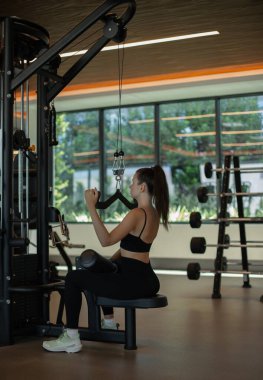 Spor salonunda makine üzerinde egzersiz yapan, dikey blok hareketi yapan, egzersiz yapan, kol ve sırt kaslarını şişiren, spor yapan güzel bir kız.