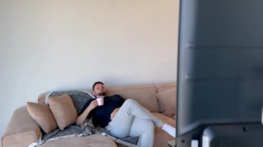 Oturma odasında yavru köpeğiyle kanepede oturan bir adam çay içip televizyon izliyor ve kanalları değiştiriyor.